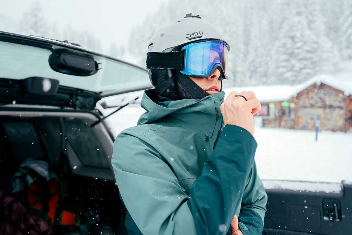 A man wearing Smith goggles and helmet zips up his jacket out in the snow