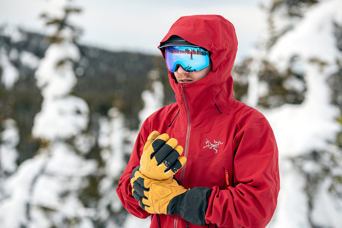 A man in a red jacket and gloves stands in the snow wearing Smith goggles.