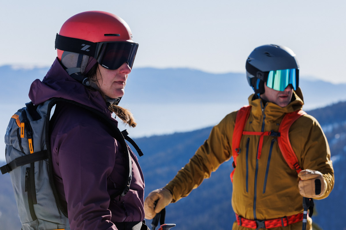 Two people standing on ski hill wearing ski helmets