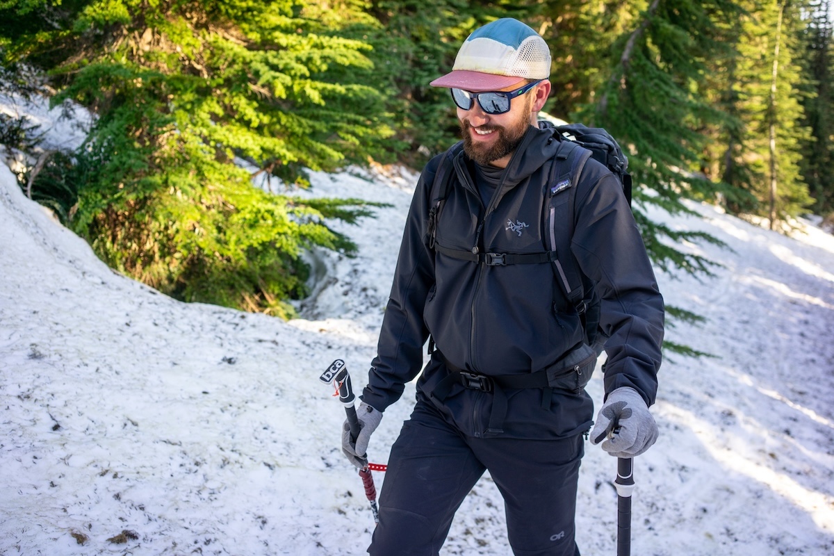 A man wearing a hat and sunglasses smiles while hiking through some snow with ski poles