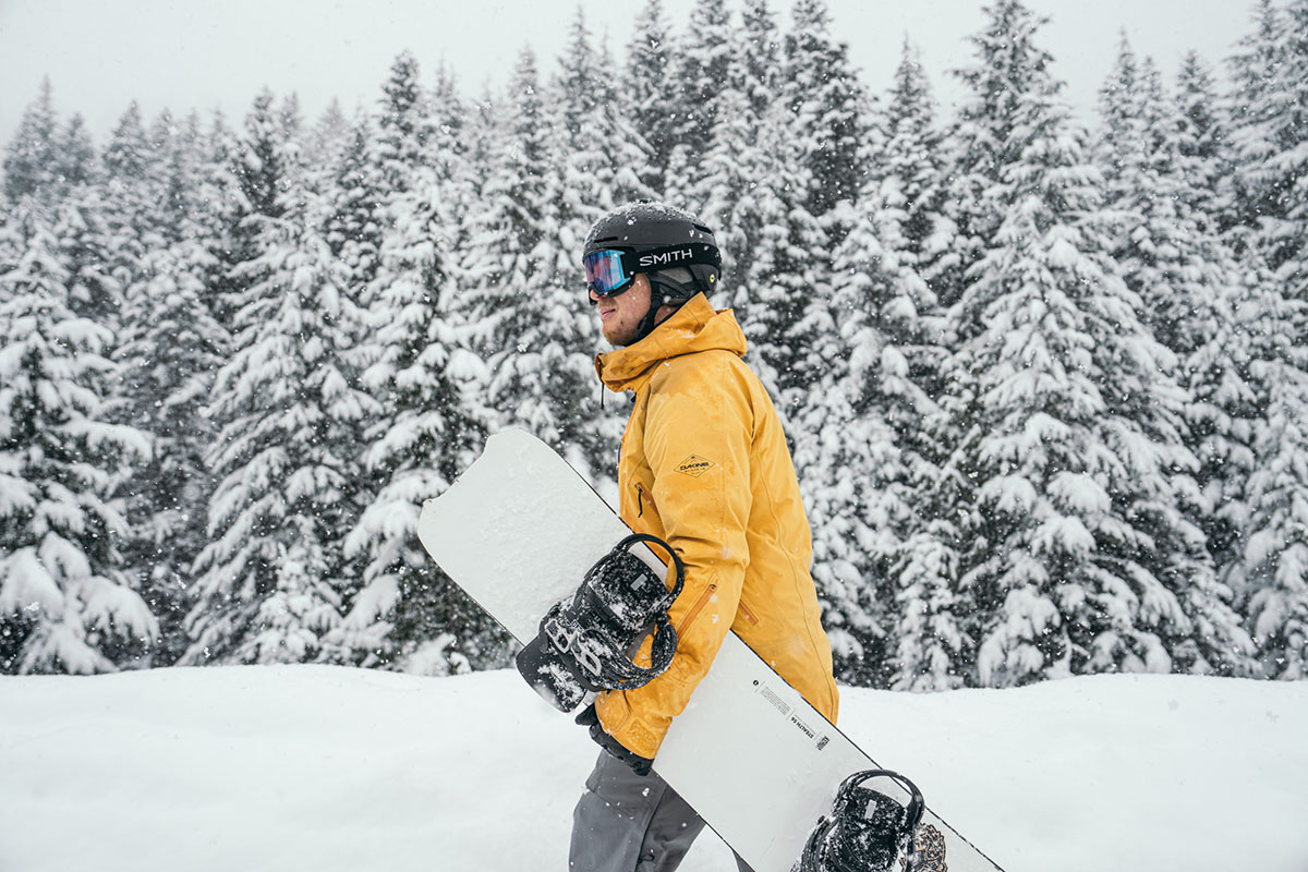 A snowboarder walking and holding his board under his arm