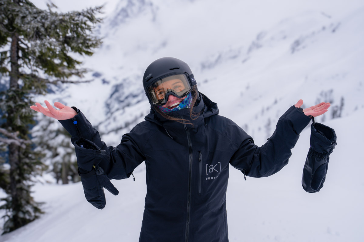 A smiling skier on a snowy slope shows off gloves dangling from wrist cuffs