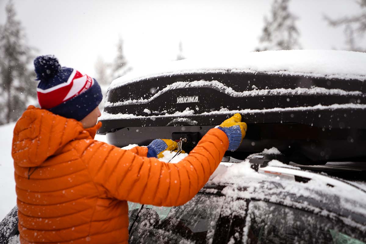 Securing a cargo roof box on top of a snowy car in an orange down jacket