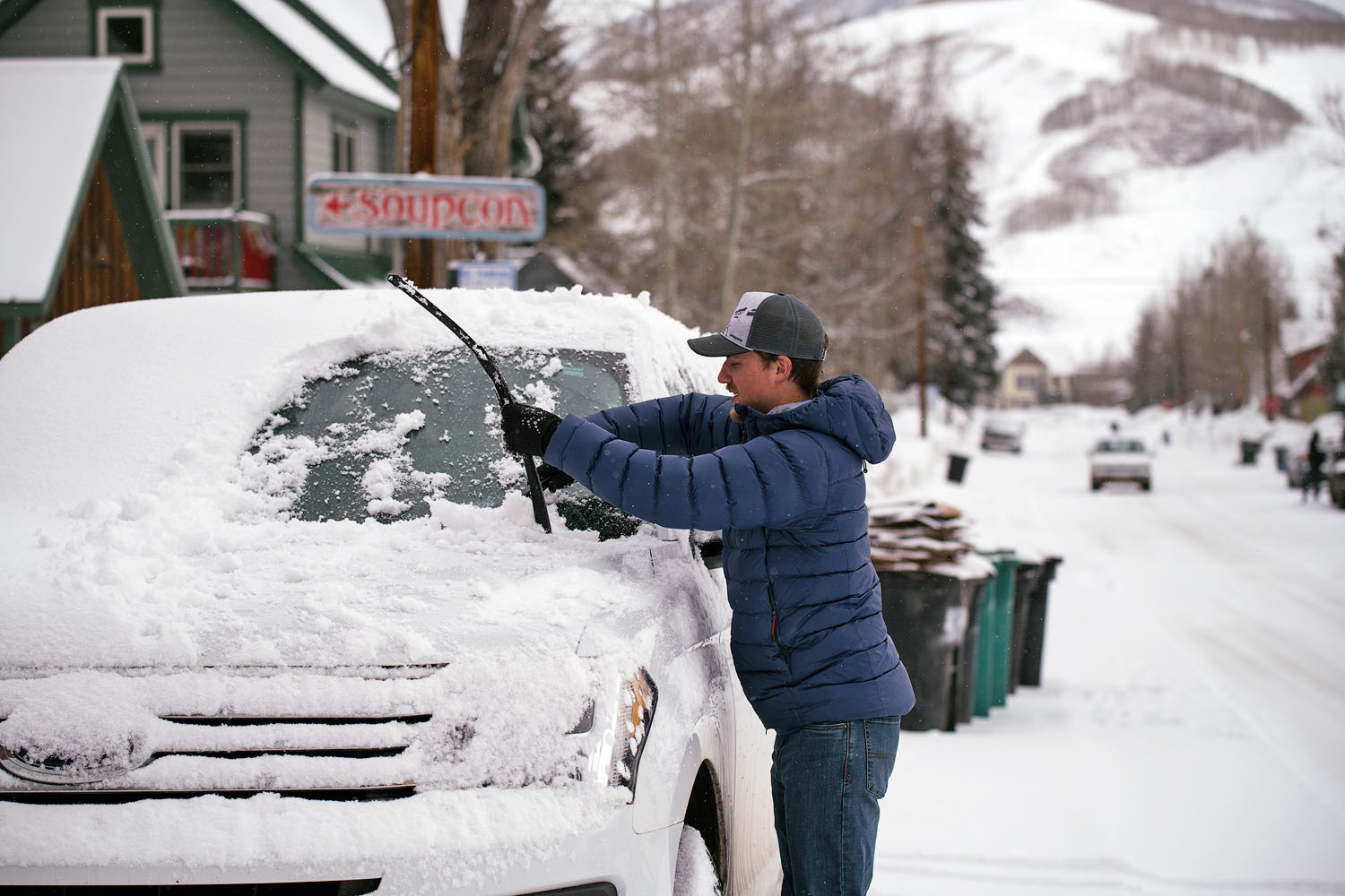A man wearing a blue down jacket cleans snow off the windshield of his car