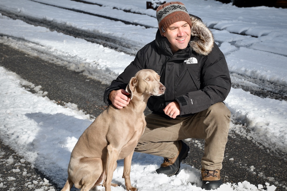A man in a black parka kneels down next to his dog on a snowy road