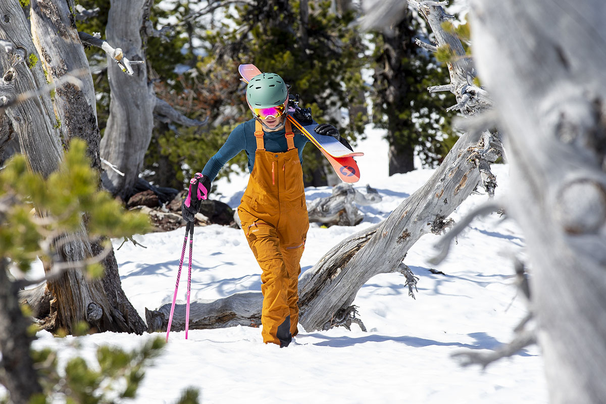 A woman in bright orange bibs and a teal base layer bootpacks up through some trees