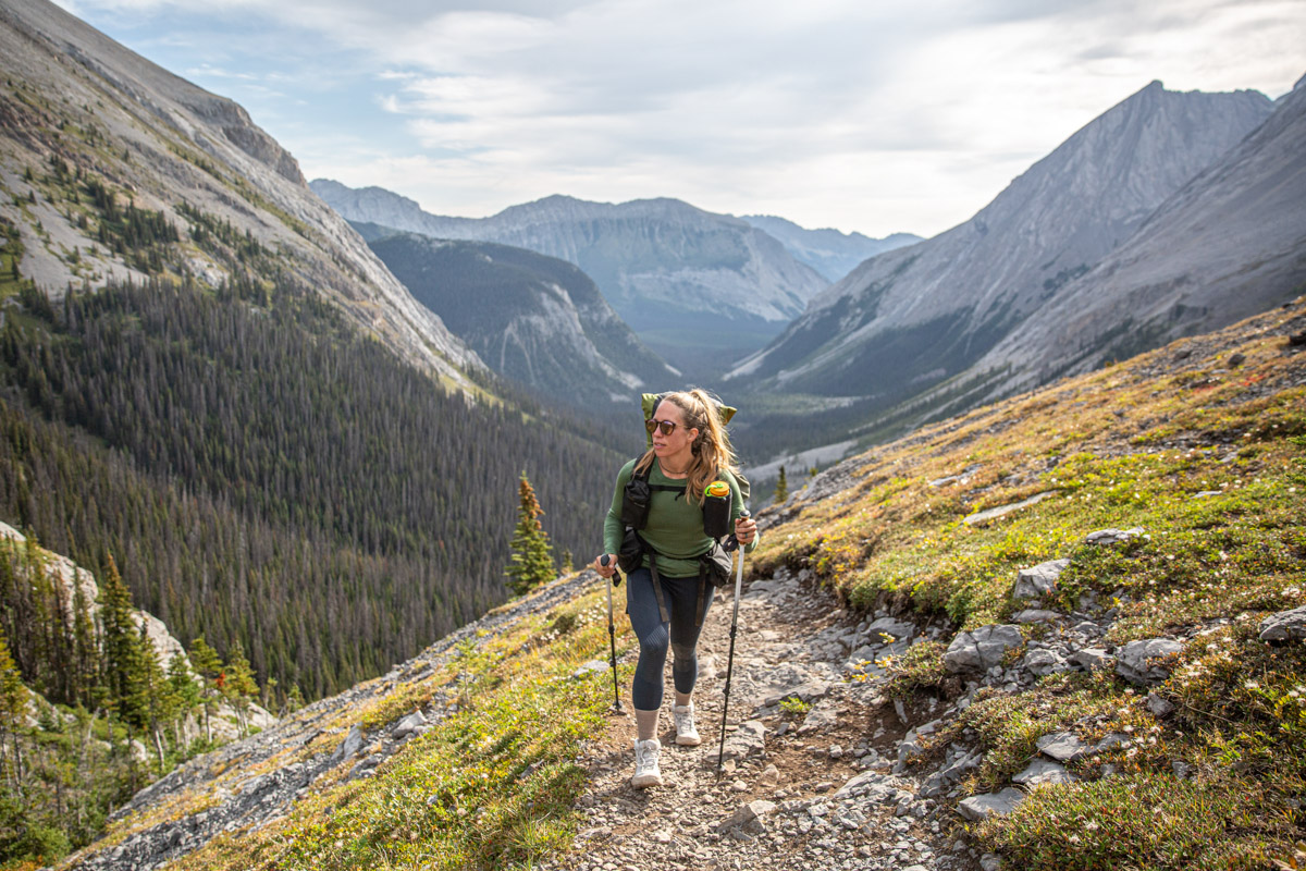 A woman in a green baselayer wears a backpack and hikes up a rocky trail with trekking poles
