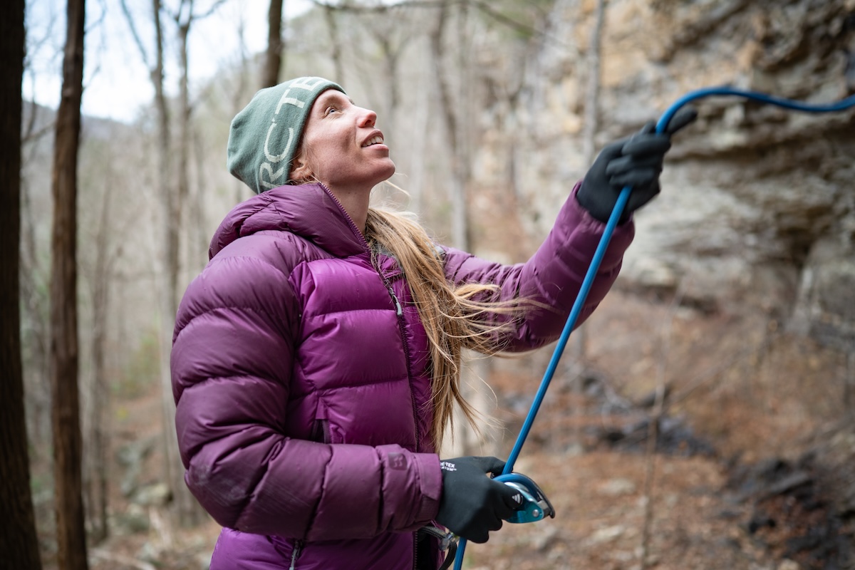 A woman in a large down jacket smiles while belaying a climber