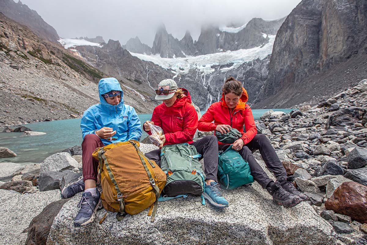Three women sit on a rock in the mountains and are opening their packs