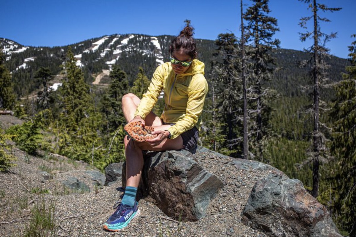 A woman sitting on a rock examines the outsole of her hiking shoe.