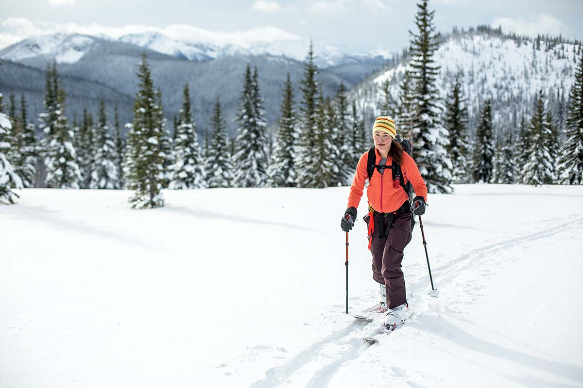 A woman ski tours toward the camera with snowy pine trees in the background