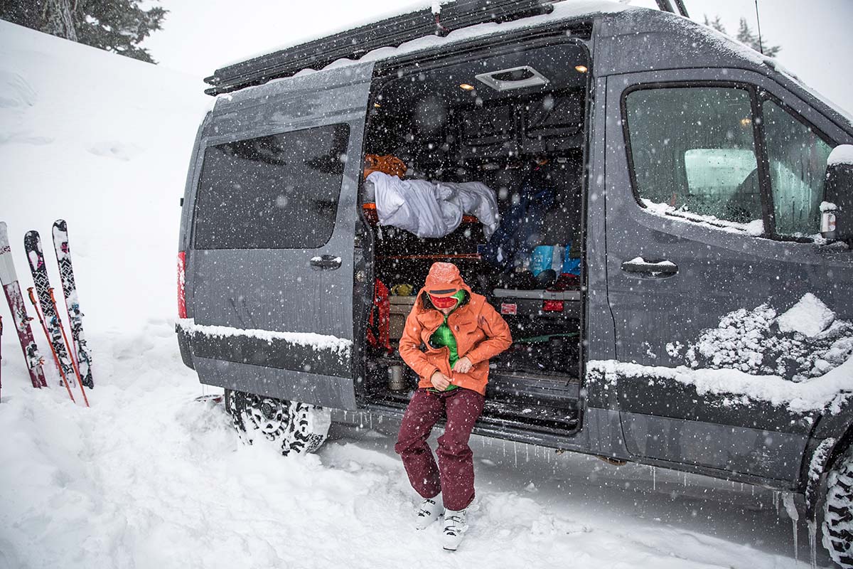 A woman sits on the edge of an open van in the snow, zipping up an orange jacket