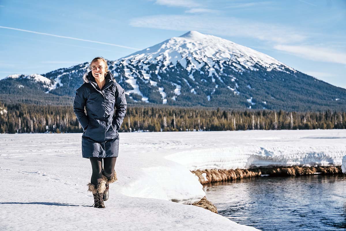 A woman in a parka and snow boots walks through the snow next to a body of water