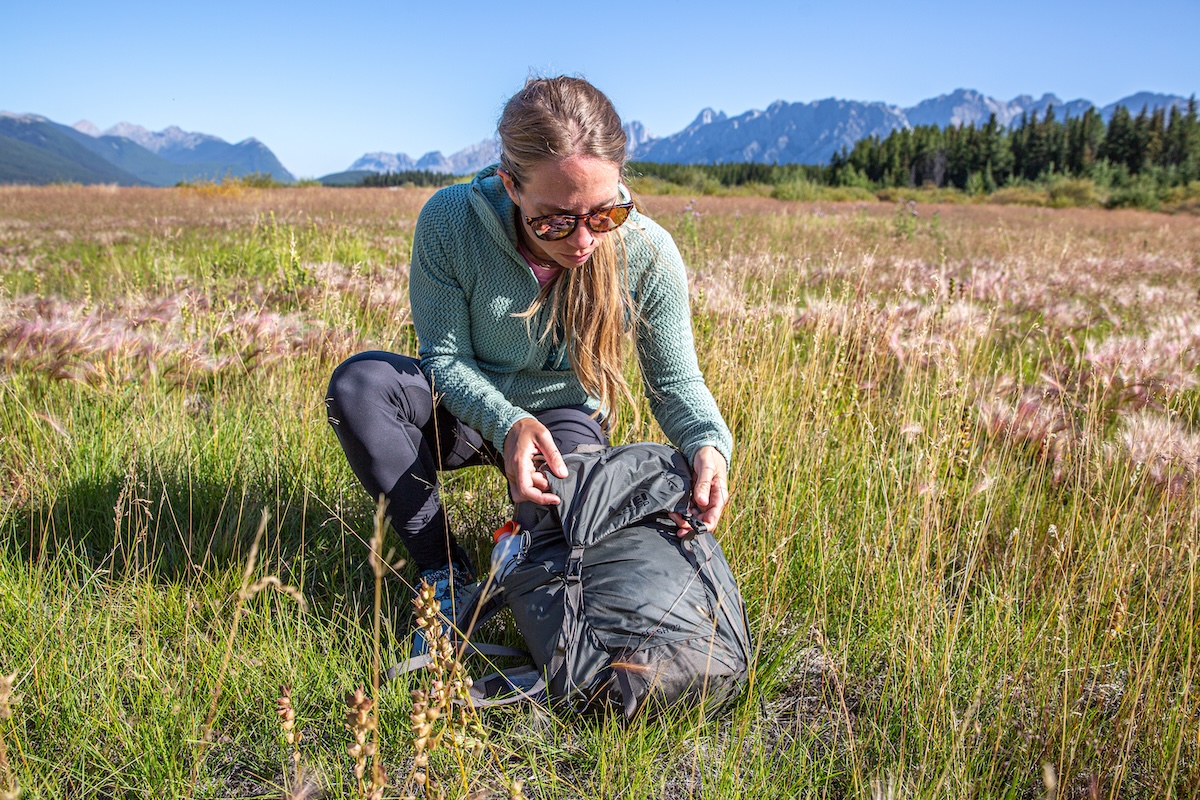 A woman in a fleece jacket kneels down in a field to adjust her backpack