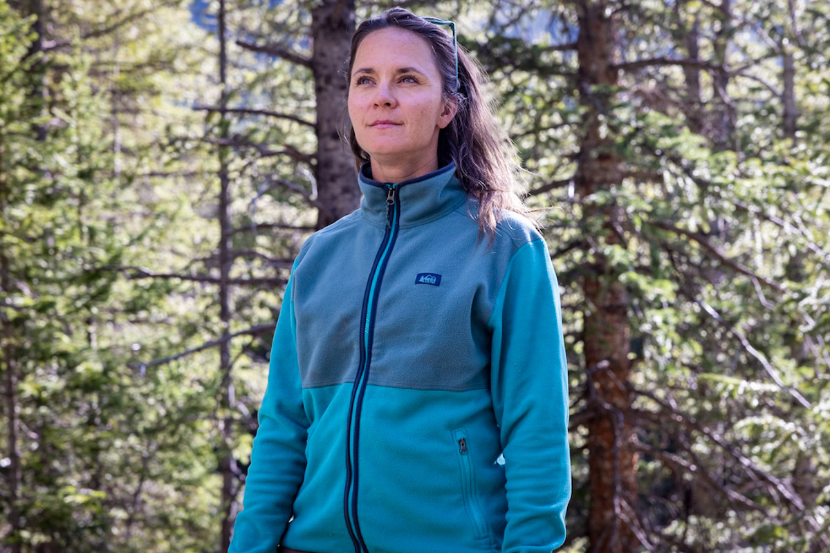 A woman in the forest wearing a blue REI zip up fleece jacket