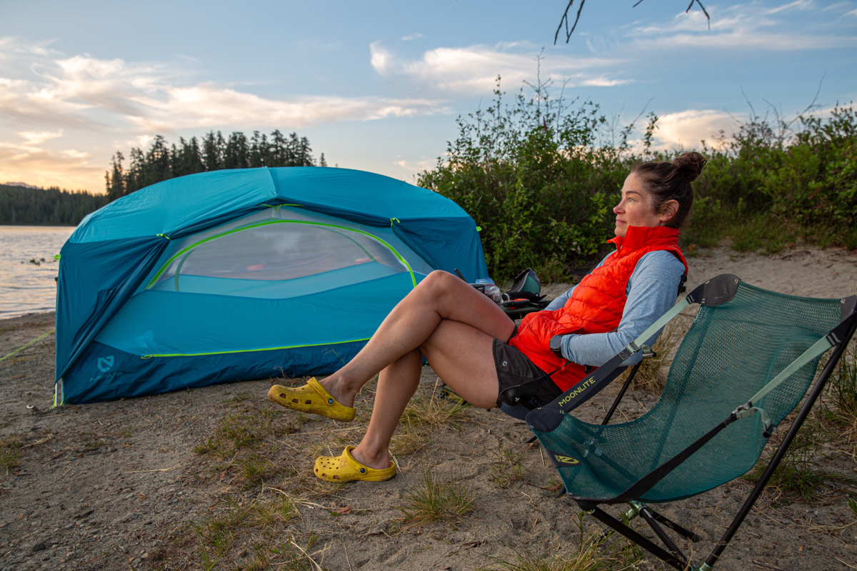 A woman sitting in a camping chair next to a blue tent and a river