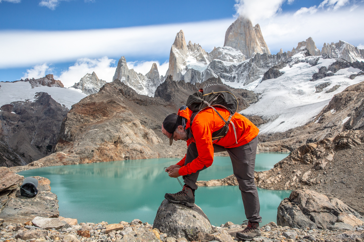 A man tying his hiking boots on a rock in front of an alpine lake and snowy mountain