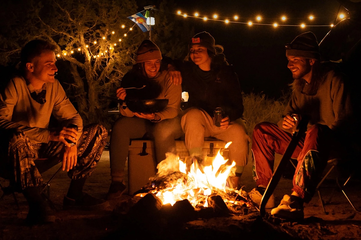 A group of campers sit around a campfire with string lights in the background