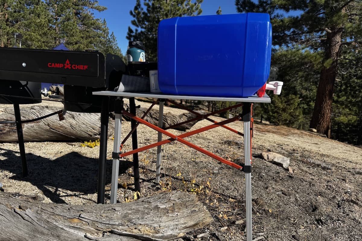 A camping table, water container, and large freestanding stove are set up in the forest