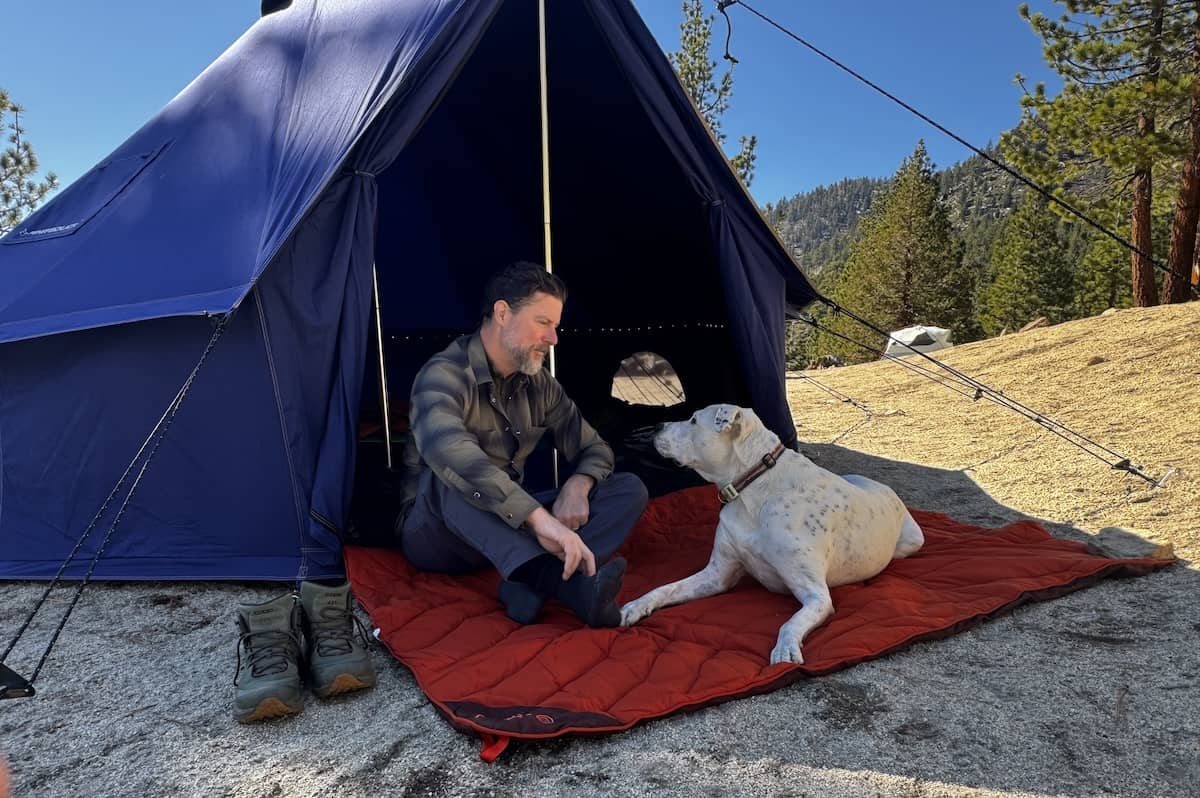 A man and a dog sit outside a blue tent on a red ground mat