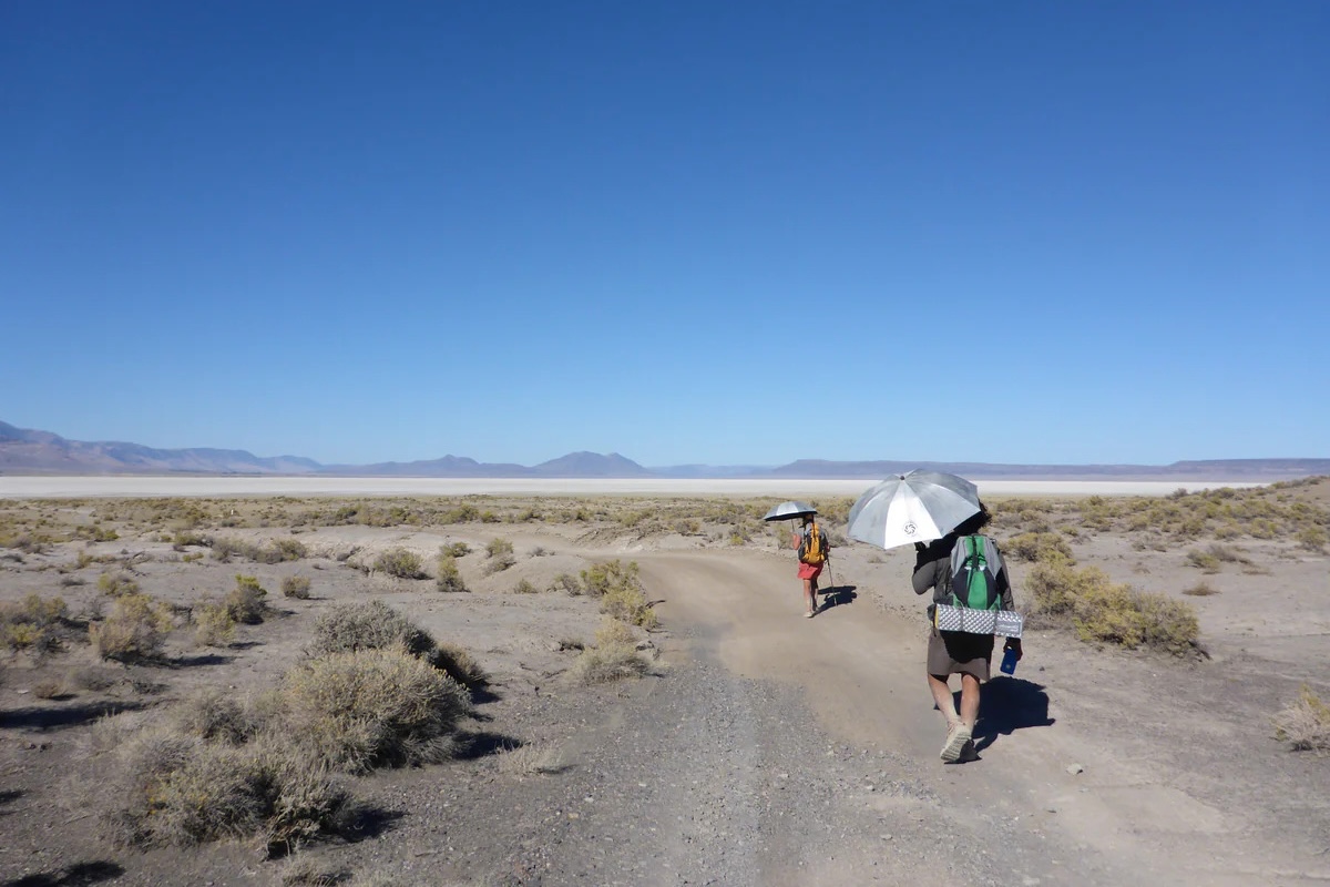 Two hikers walk in a sunny and exposed area with silver umbrellas