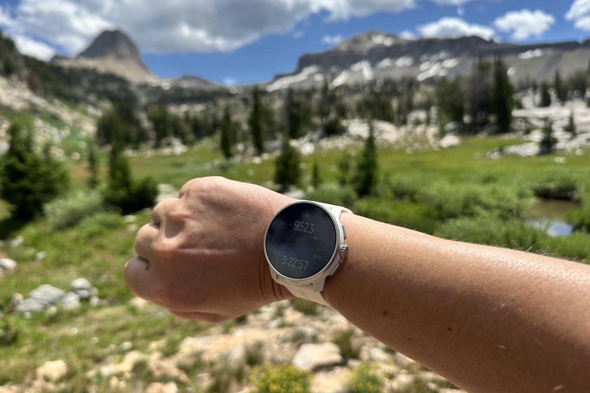 A woman's arm is shown with a smartwatch displaying the features against mountain scenery 