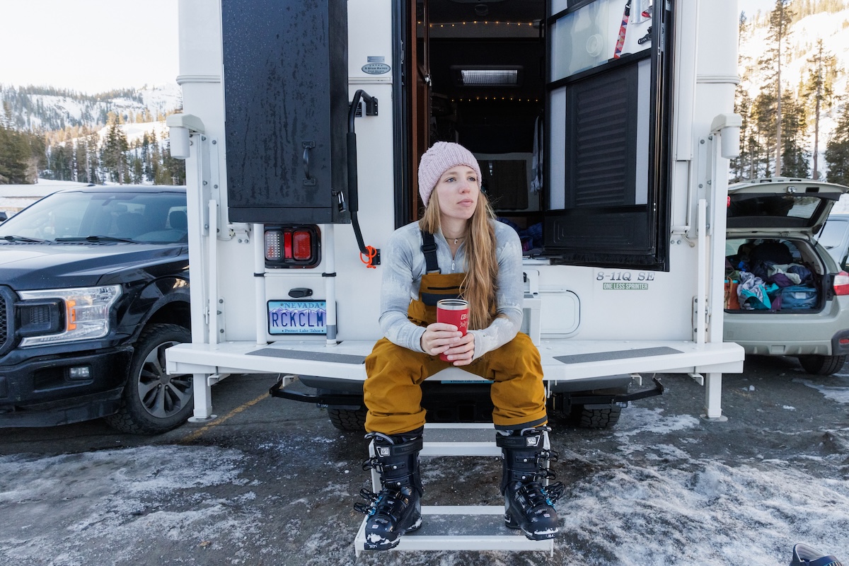 A woman in ski boots and bibs sits on the back steps of a truck camper