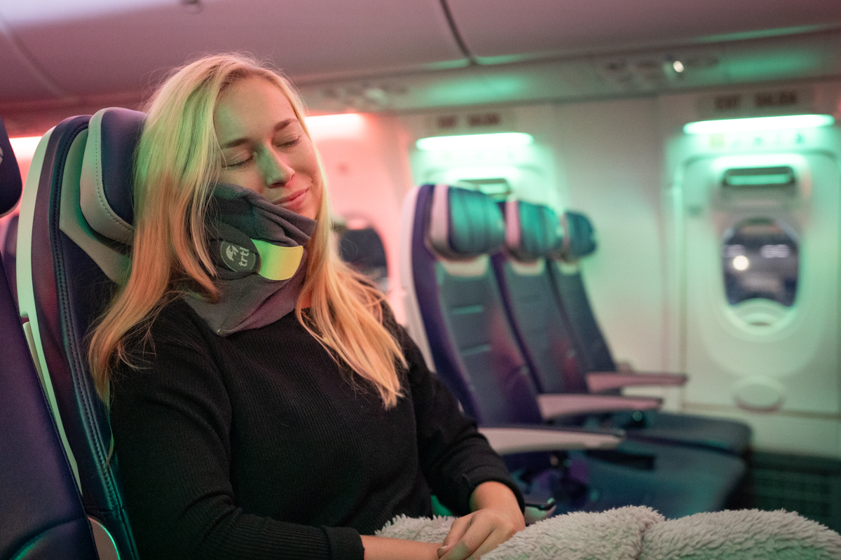 A woman sleeps in a plane seat while using the trtl travel neck pillow.