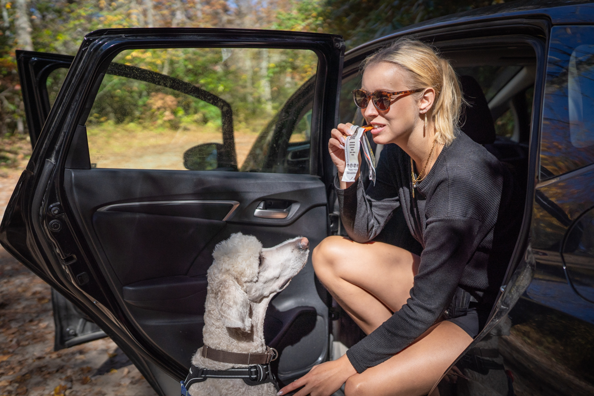 A woman sits next to her car with her dog, having a snack.
