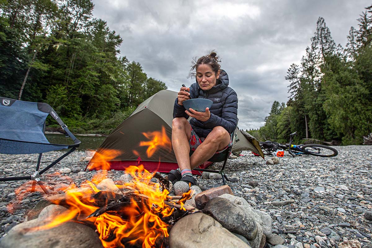 A woman sits by a fire with the Arc'teryx Cerium Hoody