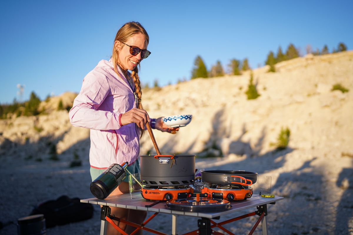 A woman smiles and serves herself food from a large pot on a camping stove
