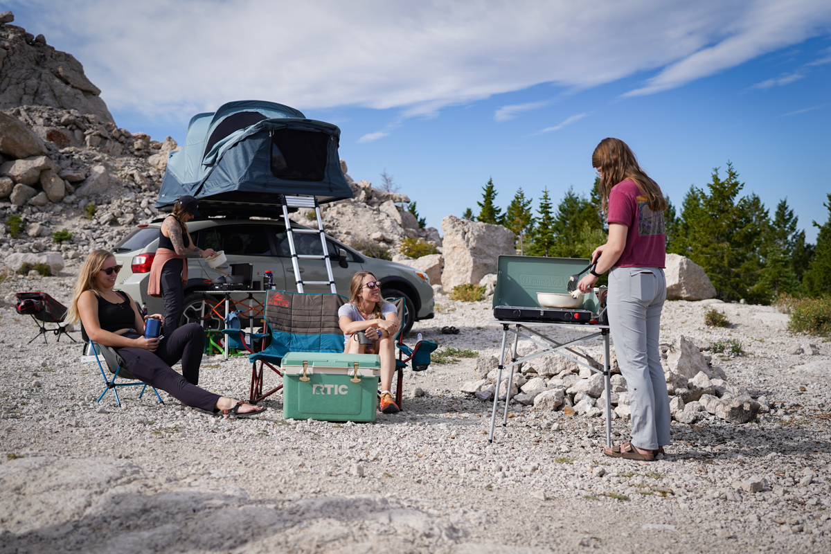 A group of campers smile and chat while some prepare food on camping stoves