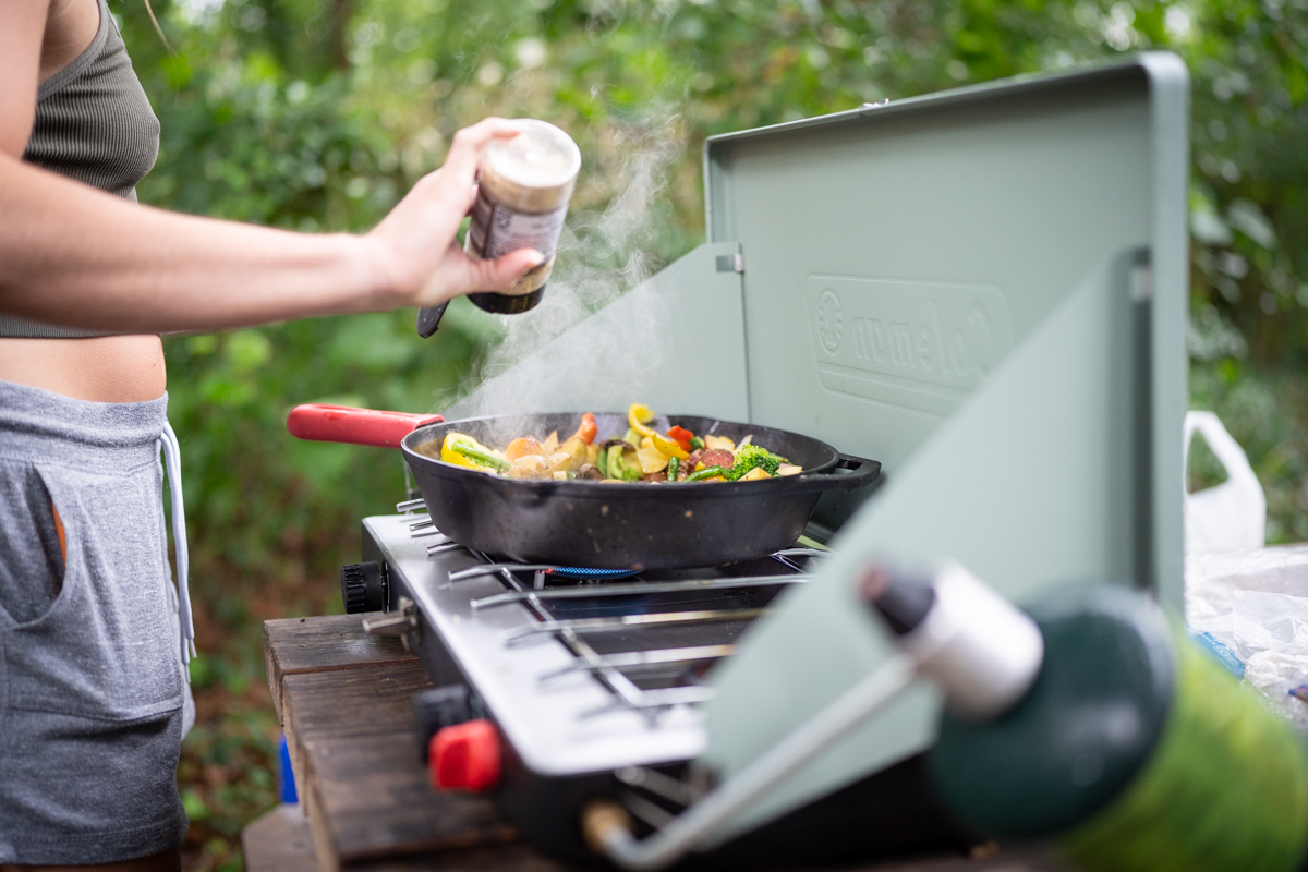 A close-up of seasoning being sprinkled on a pan filled with food on a camp stove