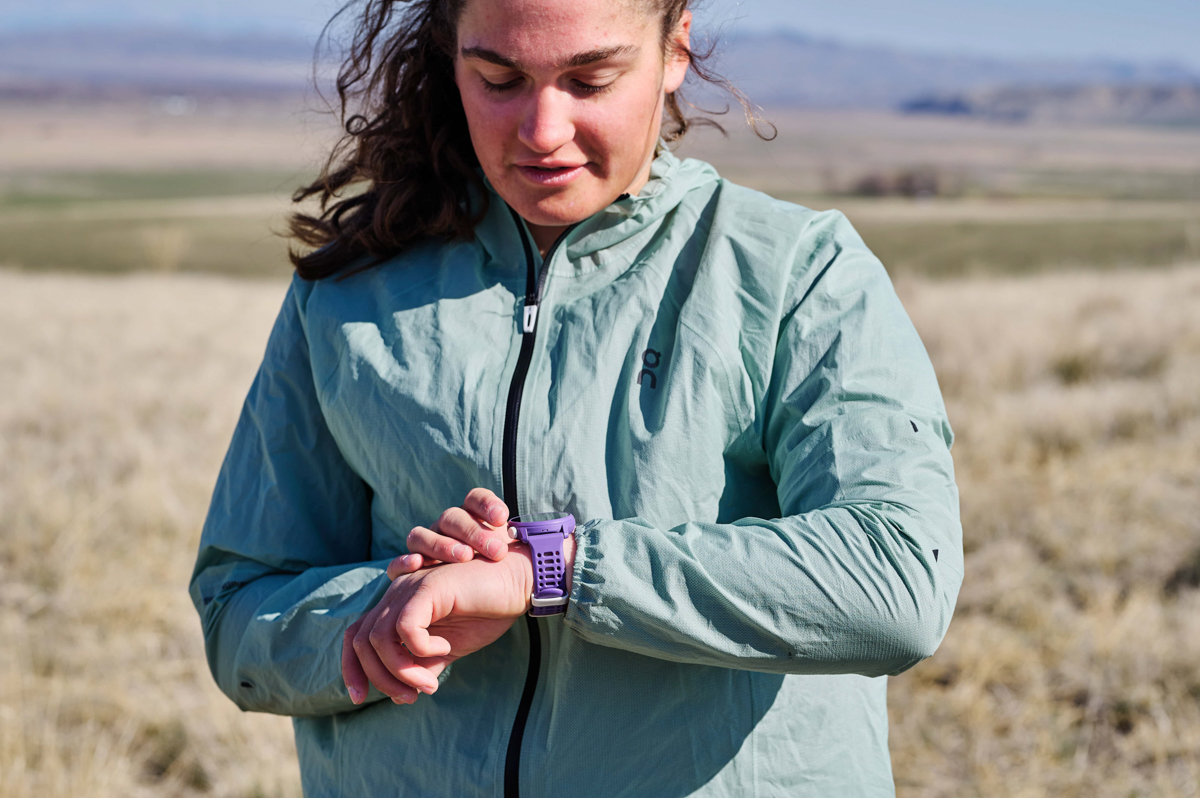 A woman looking down at her purple fitness watch while we stands in a grassy field
