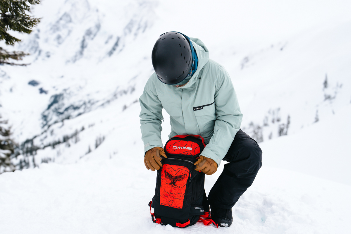 A man kneeling in the snow to unzip a pocket on a red ski backpack