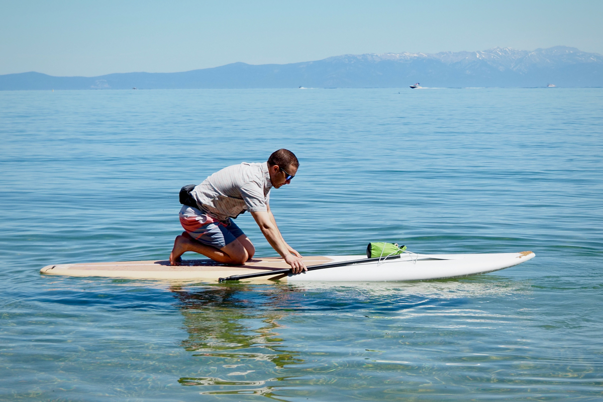 A man pulling a paddle out of the bungee on the front of a paddle board