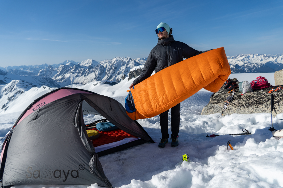 A mountaineer shows off their feathered friends sleeping bag on a snowy summit campground.