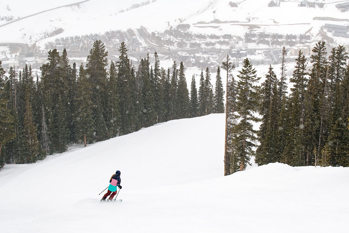 A woman is seen from far away skiing down a snowy slop between trees and with mountains in the background