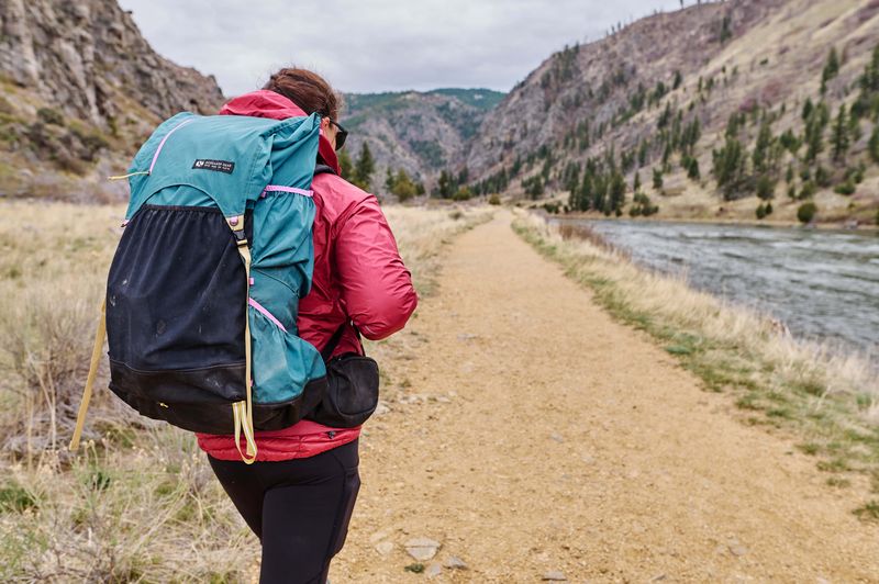 A woman walks along a path next to a river wearing a teal backpack