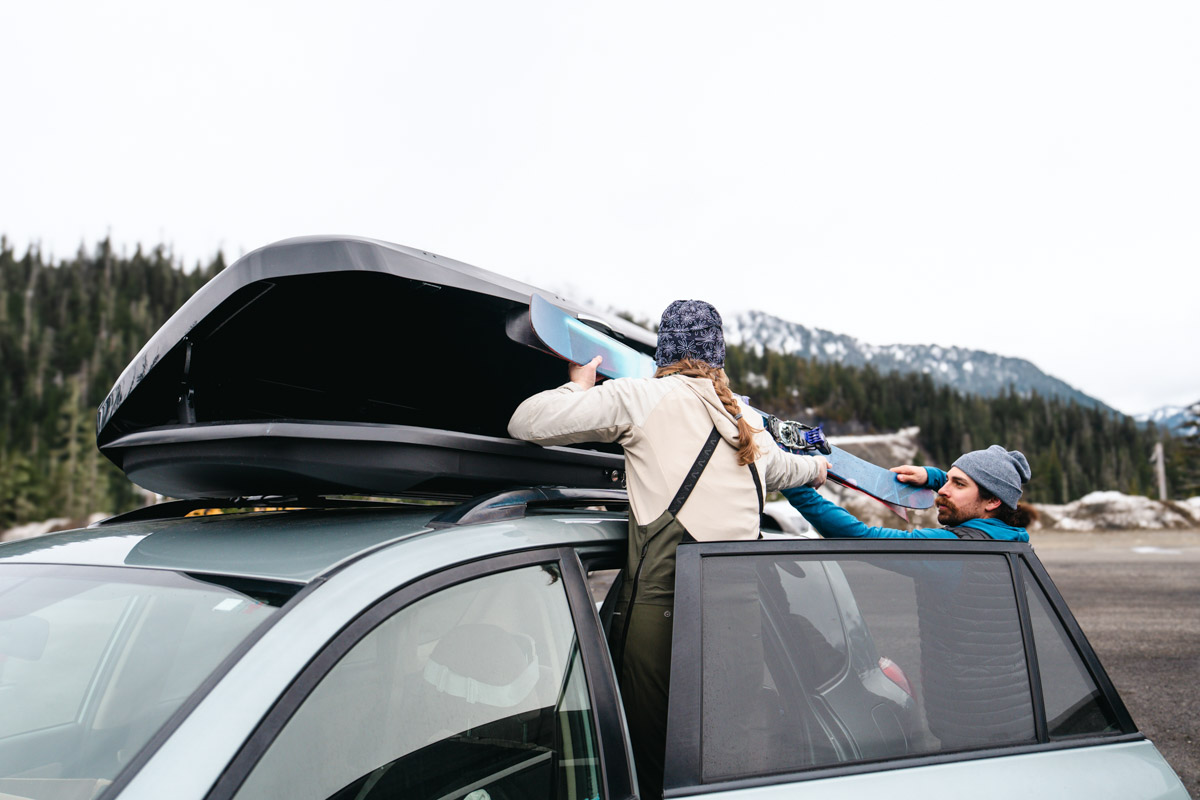 Two people lift a snowboard into a rooftop cargo box on top of a vehicle
