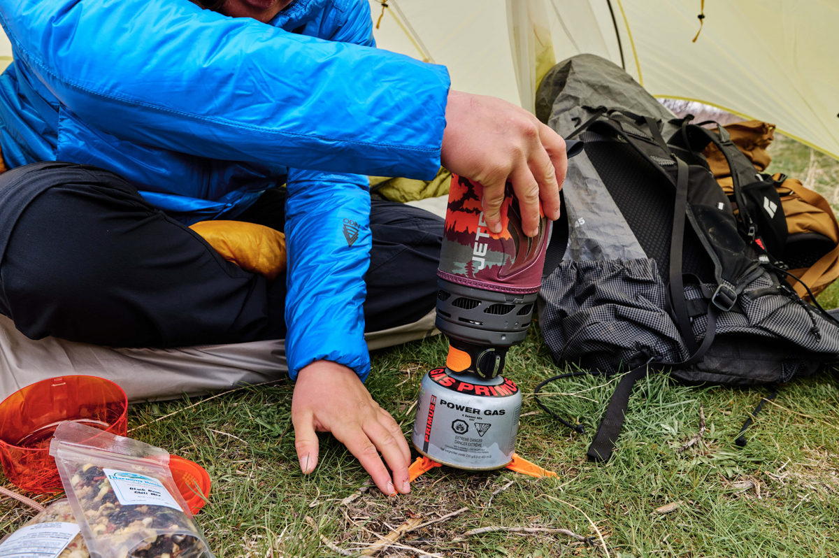 A pair of hands adjusting a backpacking stove on the ground outside of a tent