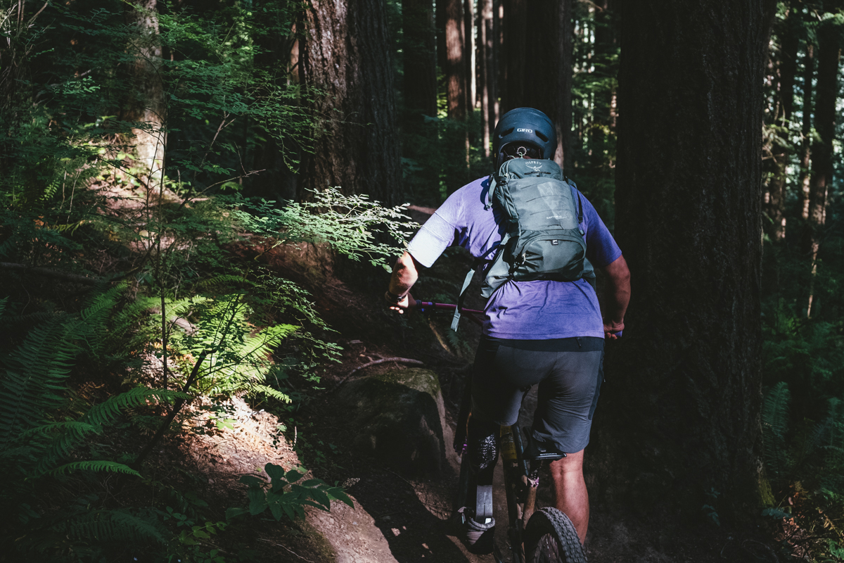 A man riding a bike through a shady forest with a green mountain bike backpack