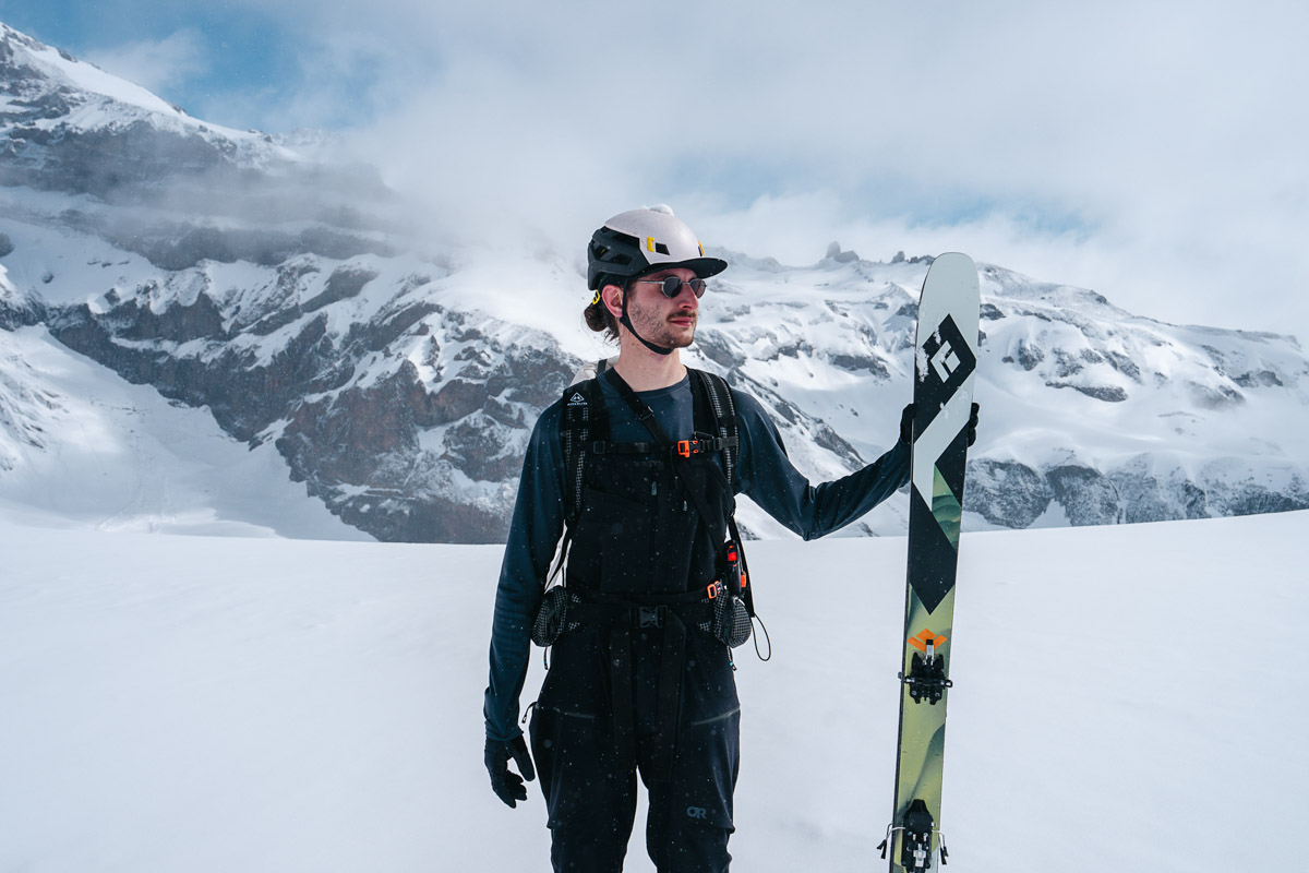 A man stands next to his skis with clouds and snowy mountains behind him