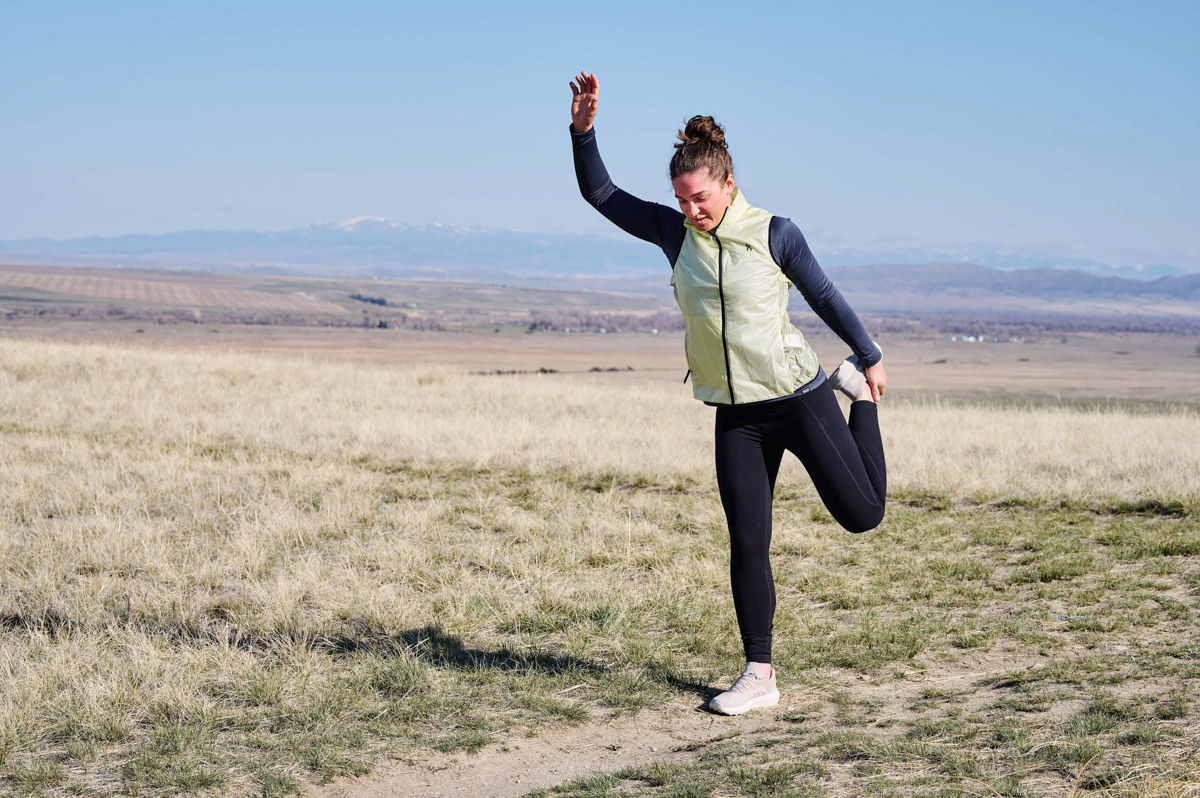 A woman in running shoes stops to stretch on a trail in a wide open meadow