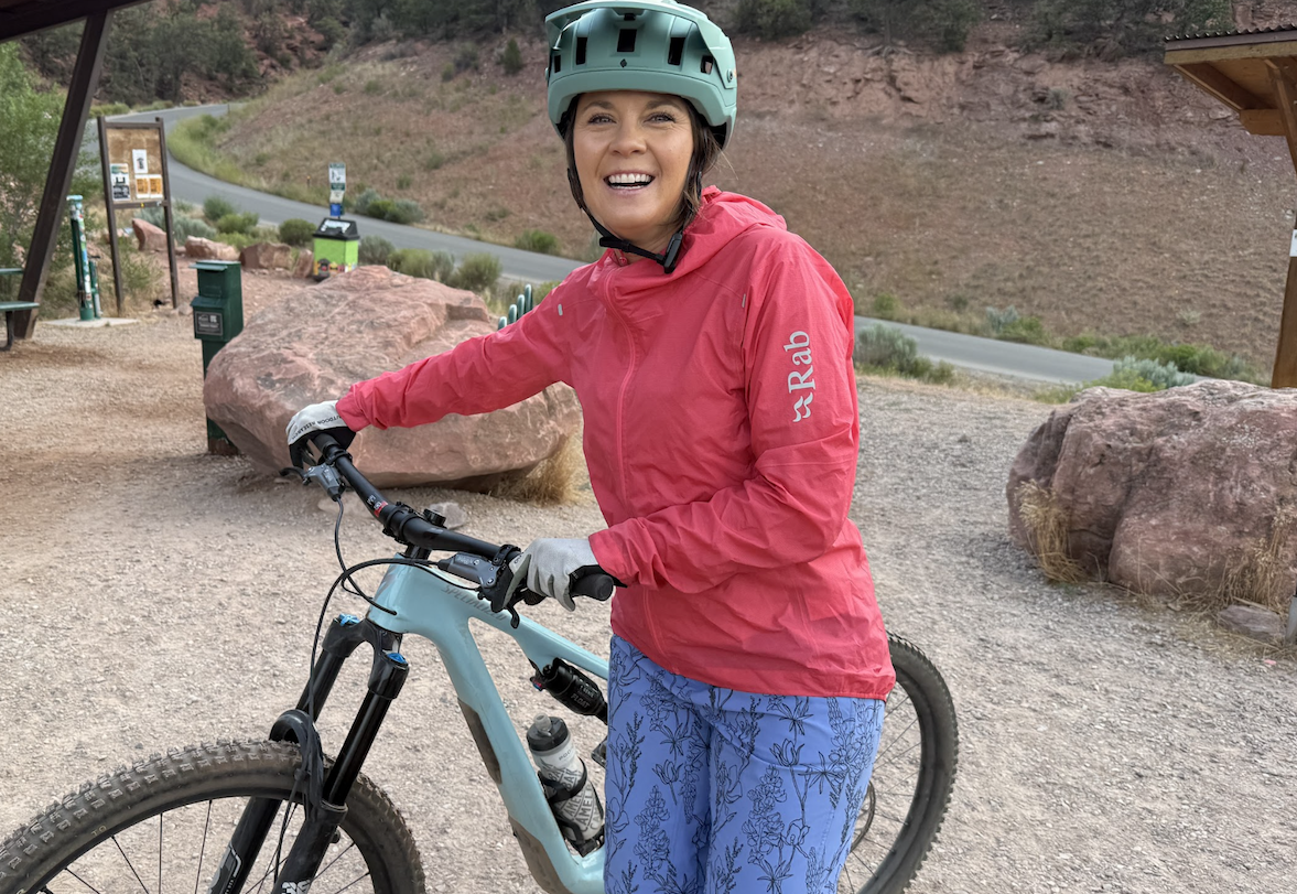 a woman smiles wheeling her bike in a dirt lot