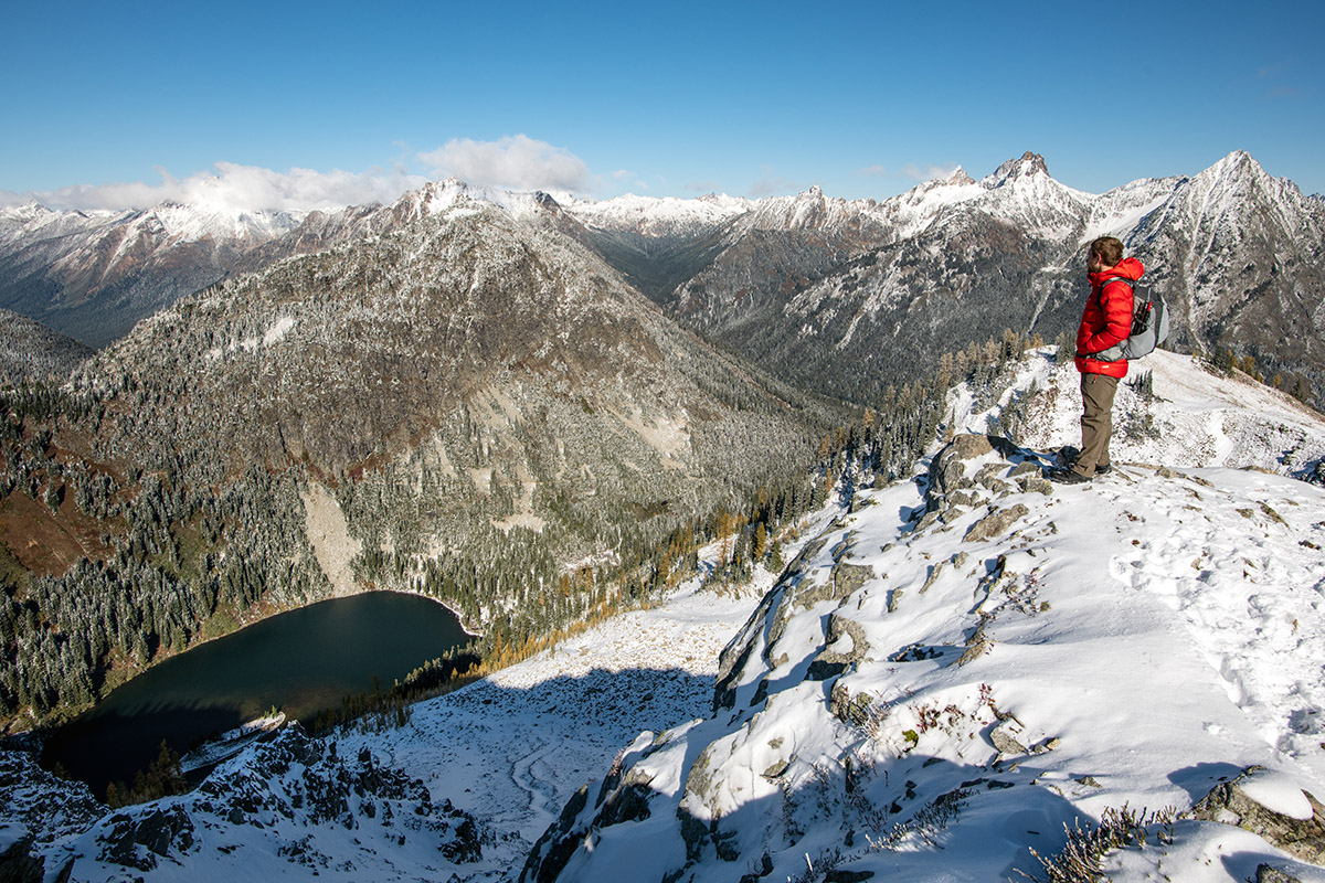 A man in a red down jacket stands on a snowy precipice, taking in the view of a surrounding mountain range