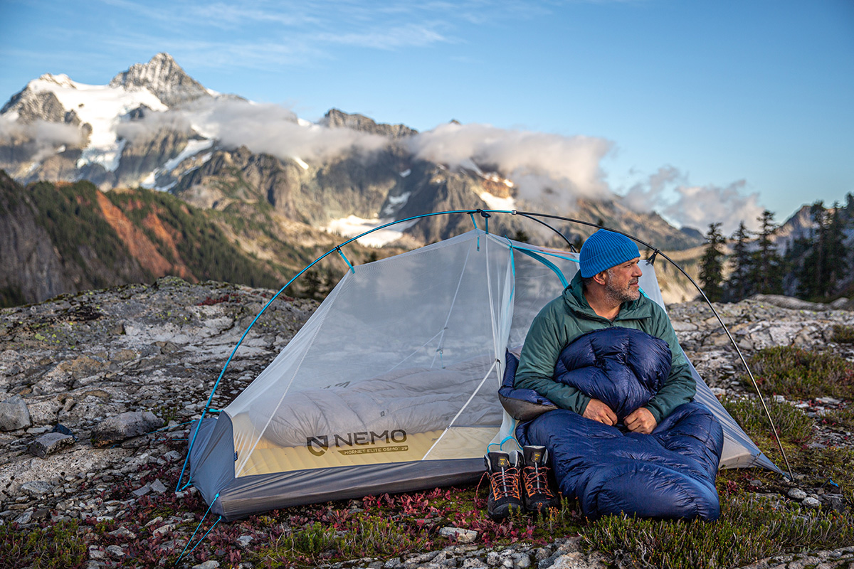 A man in a sleeping bag sitting in a tent with a mountainous background