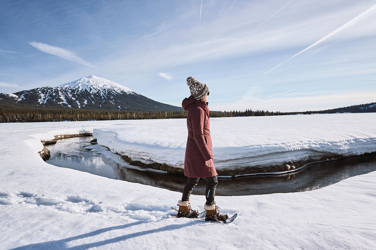A woman on snowshoes stands in front of a creek and snowy mountains