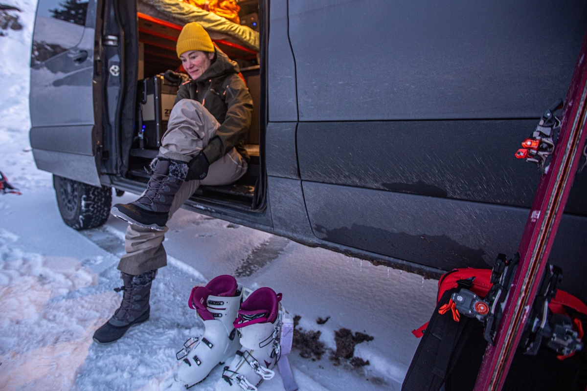 A woman pulls on a winter boot in the door to her van after skiing
