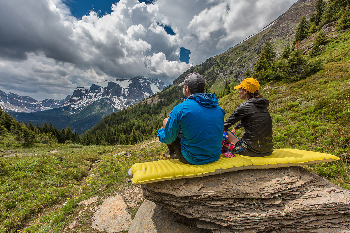 Two campers sit on an inflated mattress on a rock, while they admire the mountain view.